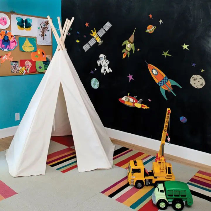 A whimsical space scene featuring colorful rockets, an astronaut, and various planets, stars, and a satellite. The SIMPLE SHAPES - OUTER SPACE REMOVEABLE WALL DECALS by SIMPLE SHAPES showcase fun designs and colors on the rockets, while the astronaut appears to be floating in a spacesuit near Earth and other celestial objects.