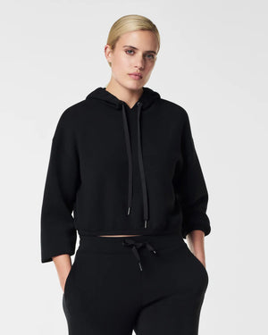 A person with short blonde hair is wearing the SPANX - AIRESSENTIALS CINCHED HOODIE in black, along with relaxed-fit matching pants. This breathable hoodie, featuring a front pocket and drawstrings for added comfort, complements the plain, light gray background as the person gazes slightly to the side.