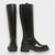 The TALL LEATHER BOOT by MICHELE LOPRIORE features two white, genuine leather knee-high boots with black soles and black stitching accents. The boot on the left is shown from the back, highlighting a full-length zipper, while the one on the right is presented from the side. Both minimalist-designed boots are showcased against a plain background. Made in Italy.