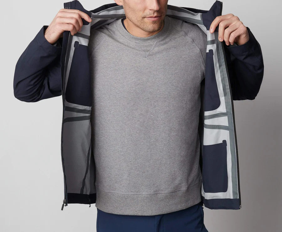 A man stands in front of a white wall wearing the MACK WELDON STORMCHASER JACKET with adjustable hood, paired with a gray shirt, blue pants, and black sneakers. He looks to the side while holding the jacket&#39;s zipper.