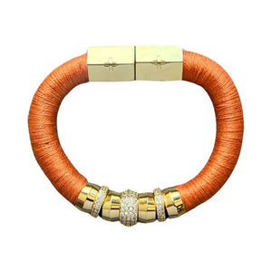 The HOLST + LEE - LUXE CLASSIC BRACELET by HOLST & LEE features an elegant orange cord with gold-plated accents, including a rectangular gold clasp adorned with several gold and diamond-encrusted beads near the clasp.