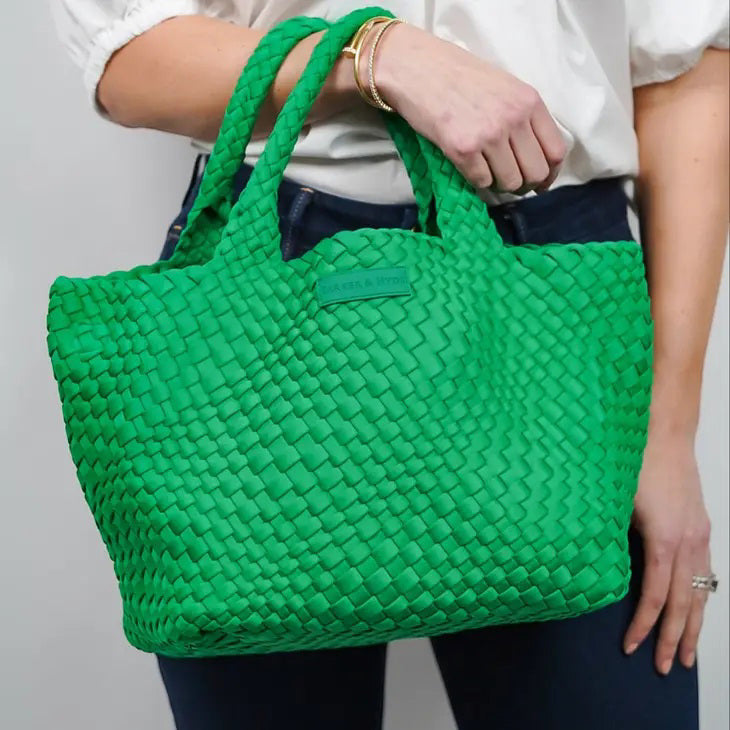 A person wearing a white shirt and dark pants holds the PARKER AND HYDE - WOVEN TOTE BAG with two handles. Their arm, adorned with a gold bracelet, stands out against a plain, light-colored background.