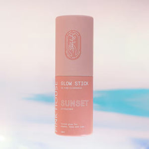 A cylindrical pink glow stick makeup product, with its cap off, is displayed against a blurred pastel pink and blue background. The label reads "PINK HOUSE ORGANICS - GLOW STICK SUNSET," indicating it’s suitable for lips, cheeks, and eyes to achieve a natural glow. The cap features a printed design.