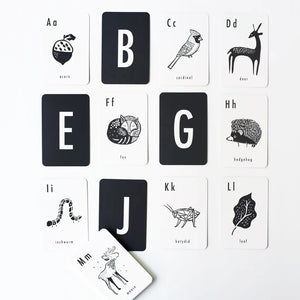 A set of WEE GALLERY - WOODLAND ALPHABET CARDS from Wee Gallery is displayed in an open white box, showcasing high-contrast illustrations of woodland animals and letters. Next to the box on a white surface lies a separate black card holder with the brand name "WEE GALLERY" and a leaf design, enhancing your child's learning experience.