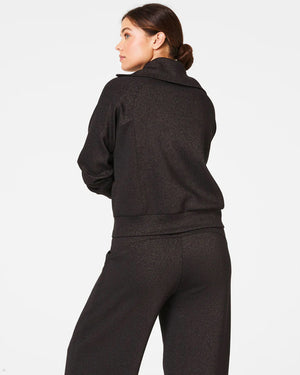 Dressed in the SPANX - AIRESSENTIALS HALF ZIP SHIMMER PULLOVER, a shimmering, gold-toned tracksuit crafted from ultra-soft fabric featuring a quarter-zip top and coordinating pants, a person stands against a simple white background with their hair styled neatly in a bun, radiating a gentle smile.