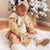 A toddler wearing a white dress, beige cardigan, and white bow sits on a fluffy white rug, playing with the SOFT SILICONE MINI ANIMAL PUZZLE by ALI + OLI that helps develop hand-eye coordination. A frosted Christmas tree is decorated in the background, adding a festive atmosphere to the cozy indoor scene.