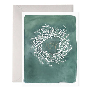 The WHITE BERRY WREATH HOLIDAY CARD by E.FRANCES PAPER features a beautifully crafted illustration of an artistic white wreath, resembling a watercolor painting, set against a green backdrop with the word "Joy" at its center. The wreath is made of delicate branches and small buds, and an envelope is subtly visible behind it.