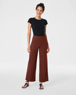 A person stands wearing a black short-sleeve top and the SPANX Stretch Twill Cropped Wide Leg Pant in brown, featuring CoreSure Tech for comfort. Paired with black sandals, they smile brightly, hands relaxed by their sides against a plain light gray background.