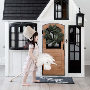 A young child in a cozy, bohemian-style bedroom holds the LITTLE UNICORN RIDER by LILY & RIVER, sparking their imagination. The room showcases children's products such as a small wooden bed with a canopy, a table and chairs, and wall decorations made of macrame. Dressed in brown with hair styled in two buns, the child enjoys playful physical activity.