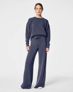 A woman stands confidently against a plain white background, wearing the SPANX Aireessentials Crew Neck Pullover and matching sweatpants made from luxurious comfort spacer fabric. She has her hair pulled back and her left hand is raised slightly. She smiles gently with her lips closed.