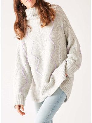 A person with long hair is wearing the MERSEA - ALPINE SWEATER by MER SEA, a loose-fitting, light grey acrylic sweater adorned with subtle purple zigzag patterns. They are also dressed in light blue jeans. The background is plain white.