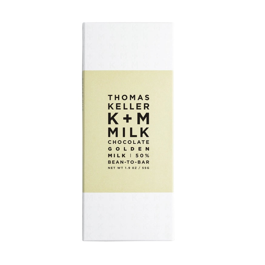 A rectangular white chocolate bar wrapper with a light beige label in the center. The text on the label reads &quot;K &amp; M EXTRAVIRGIN CHOCOLATE K+M - MILK CHOCOLATE GOLDEN MILK BAR 50% BEAN-TO-BAR NET WT 1.9 OZ / 53G.&quot; This handcrafted chocolate features flavors inspired by Golden Milk and turmeric chocolate.