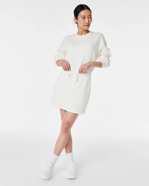 A person stands against a plain white background wearing a matching white long-sleeved sweater and the SPANX - AIRESSENTIALS SKORT by SPANX. Their hair is tied back in a low bun, and they are looking slightly to the side. They complete their look with white ankle socks and sneakers, highlighting the skort's hidden pocket feature perfectly.