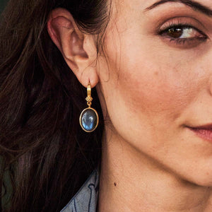 A close-up of a woman's face focusing on her left ear, which is adorned with a CAPUCINE DE WULF - CLEOPATRA OVAL DROP BLUE LABRADORITE EARRING set in 18K gold. She has long, dark hair swept behind her ear. The background is blurred.