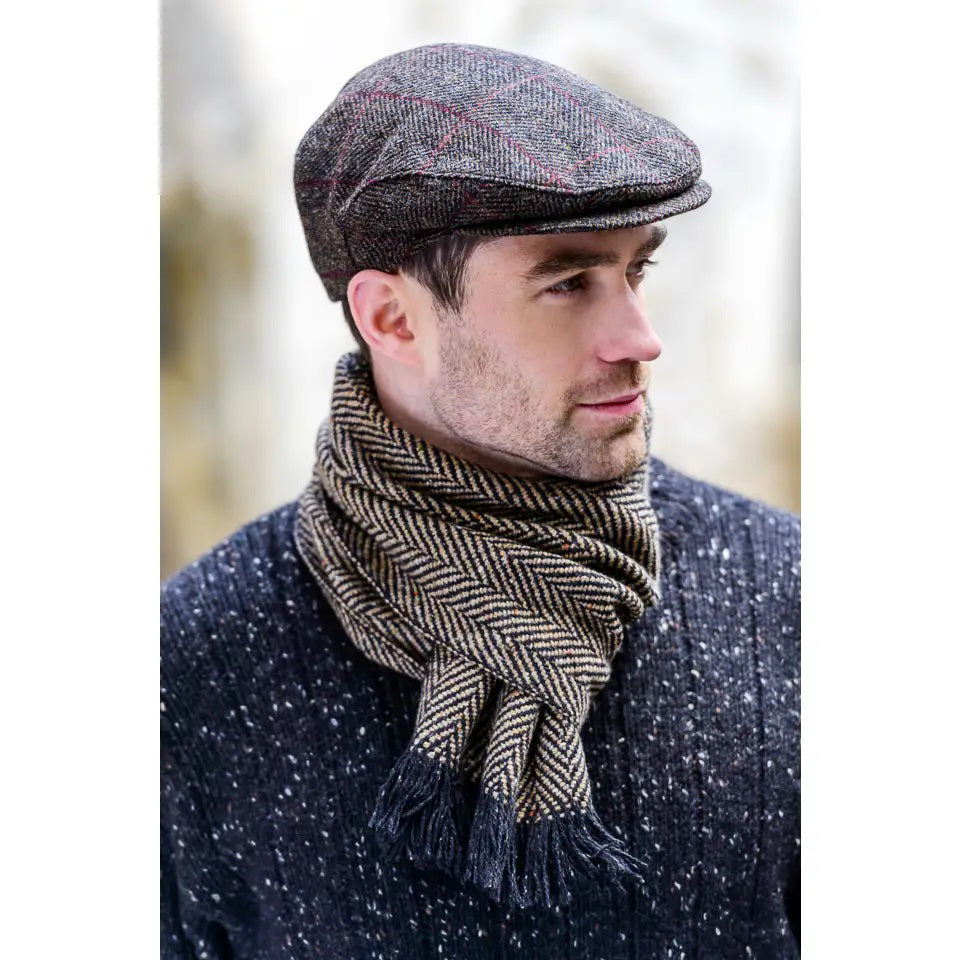 A man in a MUCROS WEAVERS - TRINITY CAP by MUCROS WEAVERS, paired with a patterned scarf and dark textured sweater, looks to the side outdoors on a cold day.