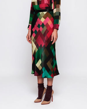 A person is wearing the MIRTO 1956 MULTICOLOR PIXEL PLAID MIDI SKIRT, which boasts a multi-colored, geometric pattern with an elastic satin waistband. They have paired it with a matching long-sleeve top, glittery ankle socks, and high-heeled shoes. The skirt features a vibrant mix of red, green, black, and beige colors that evoke a pixel print design. The background is plain white.