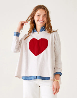 A person with long hair sits on a wooden stool against a white background, wearing an oversized MER SEA Catalina Long Sleeve Slub Tee with Heart, white pants, and blue boat shoes. They look at the camera and smile.