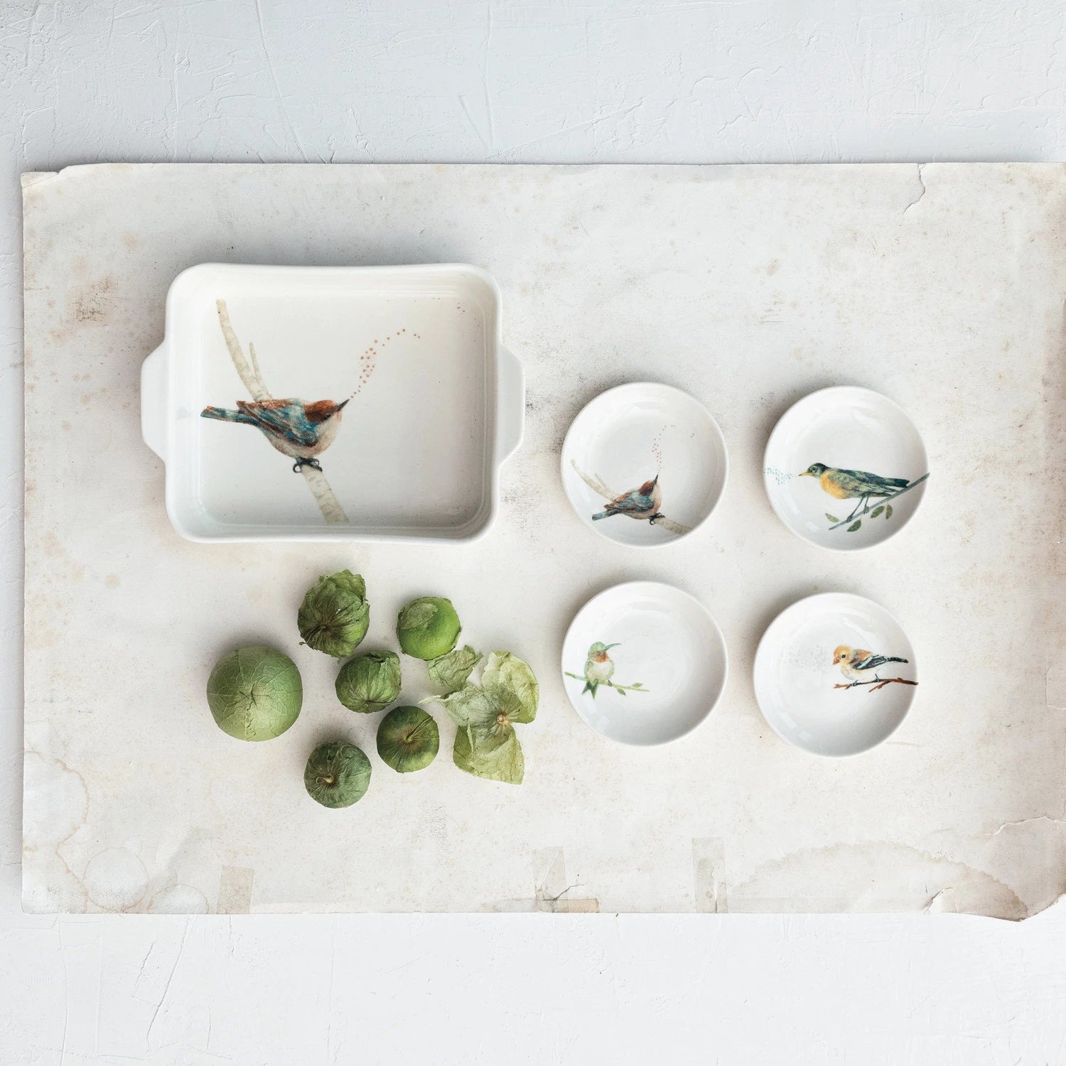 The BIRD CURIO DISH by CREATIVE COOP features a ceramic rectangular dish and four bowls with bird illustrations on parchment paper, complemented by a stoneware dish next to seven green tomatillos with husks for a rustic composition.