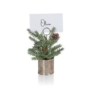 Two ZODAX PINE IN SILVER BUCKET PLACE CARD HOLDERS, featuring place cards with the names "Olivia" and "Sophia," adorn a gray surface. Adjacent to them, a lit candle in a 3 in x 5.75 in birch-patterned holder sits on a metal tray against a dark green backdrop.