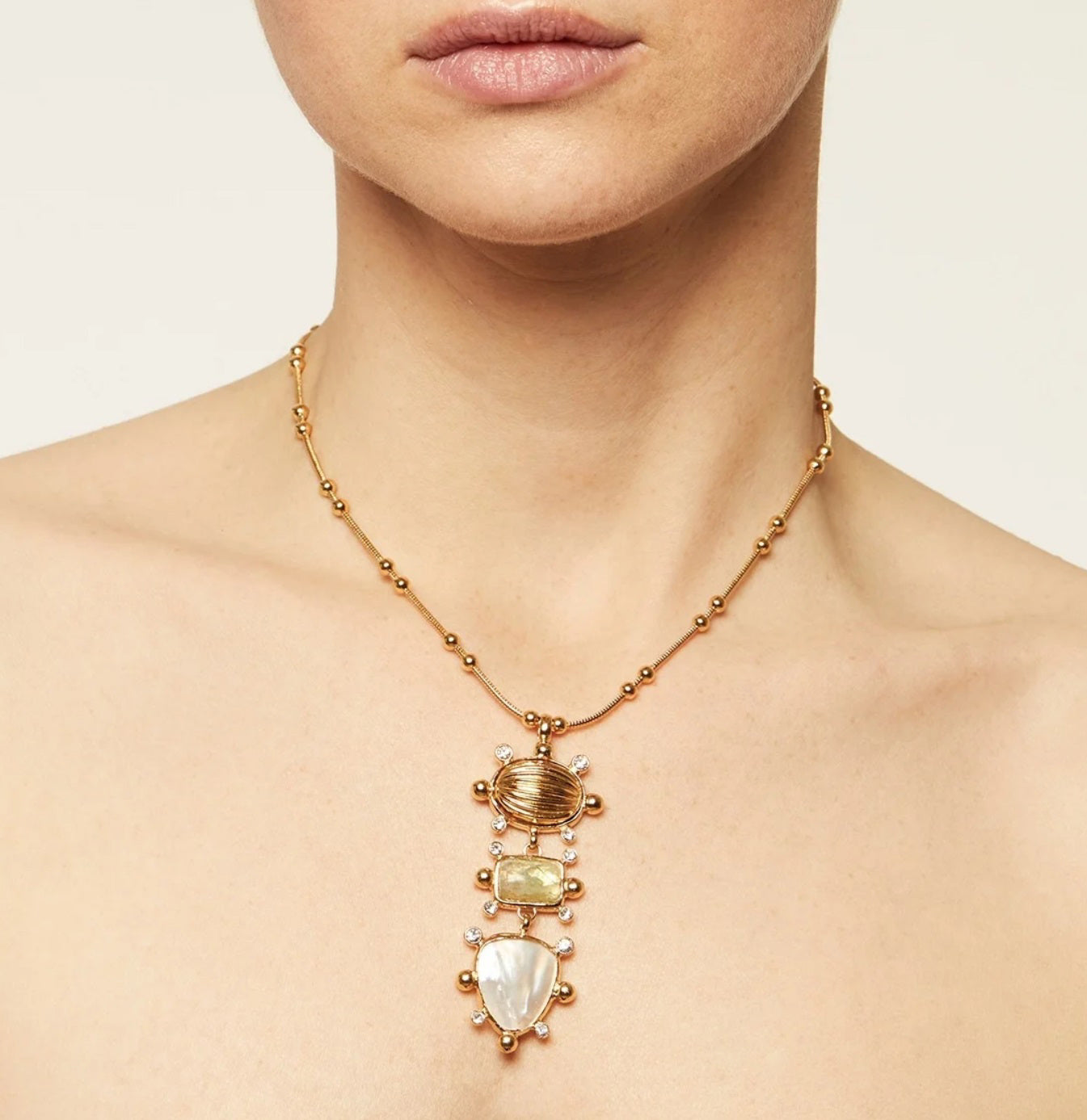 The GAS BIJOUX TIKI NECKLACE is a gold-plated necklace featuring a pendant with three gemstones in oval, rectangle, and teardrop shapes. These stones are set in a vertical arrangement within a gold frame, resembling a lucky charm, and hang from a gold chain adorned with small bead-like elements.