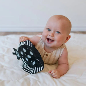 The WEE GALLERY - ORGANIC CRINKLE FISH by WEE GALLERY is a charming black, fish-shaped oven mitt made from organic cotton, featuring white polka dots scattered across its surface. It includes a handle resembling a fish tail and an opening at the side for inserting your hand. Additionally, there's a black tag attached near the middle.