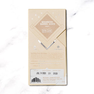 The image shows a light brown rectangular package of Ritual Chocolate labeled "RITUAL CHOCOLATE - BOURBON BARREL AGED CHOCOLATE BAR 75%." This limited edition chocolate, crafted from aged cacao nibs, features geometric designs in silver and white and contains 2.12 oz (60g) of chocolate. The background is a marble surface.