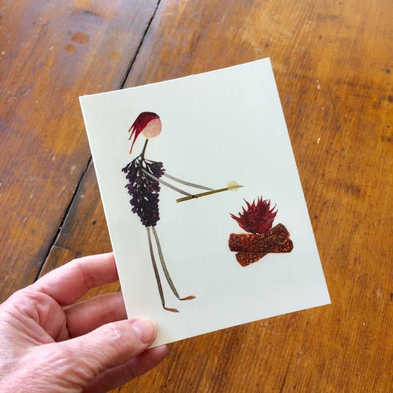 A hand holds the CAMP FIRE CARD by PETAL PEOPLE PRESS, showcasing a pressed flower notecard with a stick figure that has red hair and wears a dress made of leaves. The figure is depicted roasting a marshmallow over a small, stylized campfire, set against the backdrop of a wooden surface.