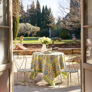 A sunlit outdoor dining scene showcases a COULEUR NATURE JARDIN BLEU & VERT TABLECLOTH 59X59, set for two. A bouquet of white roses, a wine bottle, and glasses add elegance. The garden view, framed by open French doors, features trees and shrubs in the background.