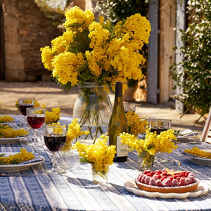 A sunlit outdoor table features the COULEUR NATURE PROVENCE AVIGNON TABLECLOTH (71x71), adorned with glasses of red and white wine, a bottle, a raspberry tart, and vases of bright yellow flowers as centerpieces, creating a vibrant rustic ambiance with French elegance.