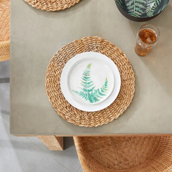 The SEAGRASS ROUND PLACEMAT from NAPA HOME AND GARDEN is made from natural straw fibers and features a braided design with varying shades of light and dark brown, creating a textured pattern. This home decor piece lies flat on a white surface and is perfect for complementing Italian ceramics.