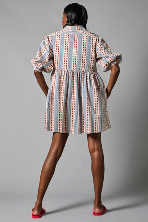 A person stands with their back to the camera, wearing the eco-friendly HOPE FOR FLOWERS TUCKED SLEEVE SHIRTDRESS, GOTS Certified with vertical stripes and dots in red, white, and blue. The dress has puffed sleeves and pairs perfectly with red open-toe sandals. The background is gray.