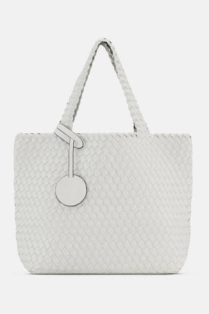 The WOVEN REVERSIBLE TOTE BAG by LINES OF DENMARK is a white tote featuring two shoulder straps and a circular faux-leather tag on one strap. This shopper showcases a textured, interlaced pattern and reversible design, providing stylish and elegant dual styling options. The image background is plain white.