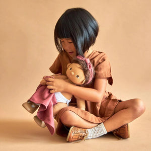 A DINKUM DOLL from OLLI ELLA USA features soft brown hair and comes dressed in a white knit romper paired with brown shoes. This charming doll has an embroidered face with brown eyes, a small nose, and a gentle smile. Its posable body makes it perfect for imaginative playtime, and it is posed standing upright.