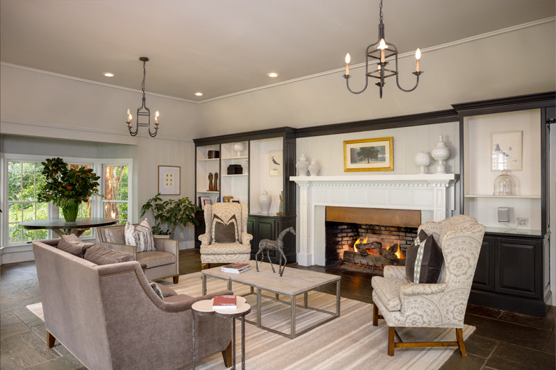 A cozy living room with a lit fireplace, gray sofas, patterned armchairs, a striped rug, two chandeliers, built-in shelves with decor, and large windows letting in natural light.