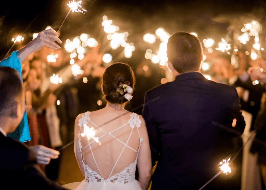 A bride and groom walk together at night, surrounded by guests holding sparklers. The bride wears a dress with a detailed back and floral hairpiece. The scene is festive and warmly lit by the sparklers.