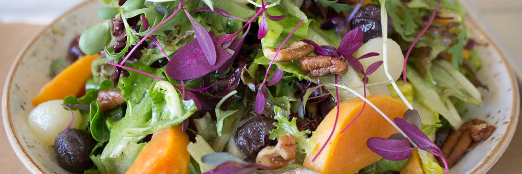 A colorful salad with mixed greens, microgreens, purple leaves, pecans, orange squash pieces, pearl onions, and dark grapes served in a bowl.