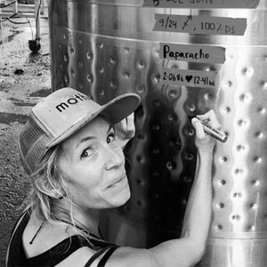 In an industrial California wine workshop, a cap-wearing individual smiles while using a marker to note the latest batch on a metal tank labeled "Paparacho" and "9/24", evoking the craft involved in A Tribute to Grace Wine Dinner by FEARRINGTON VILLAGE.