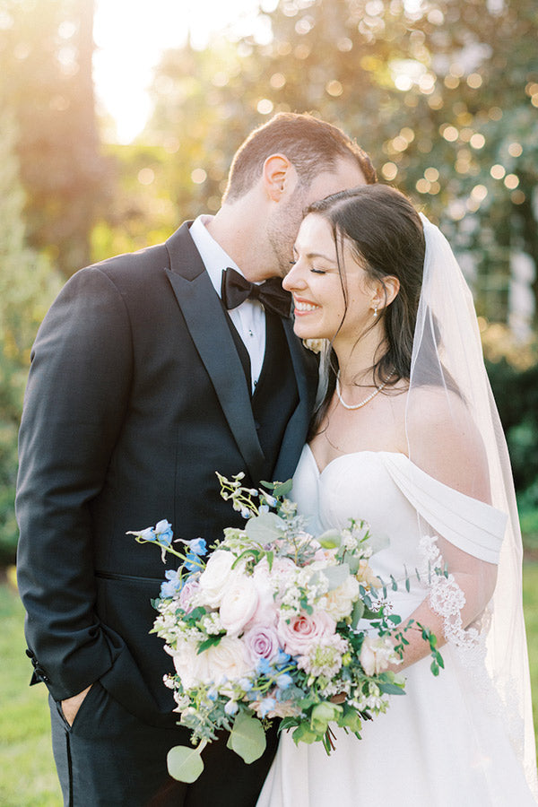 A groom in a black tuxedo kisses the bride's temple as she smiles, holding a bouquet of white and pastel flowers. The bride wears an off-the-shoulder white gown and veil. Sunlight filters through trees in the background.
