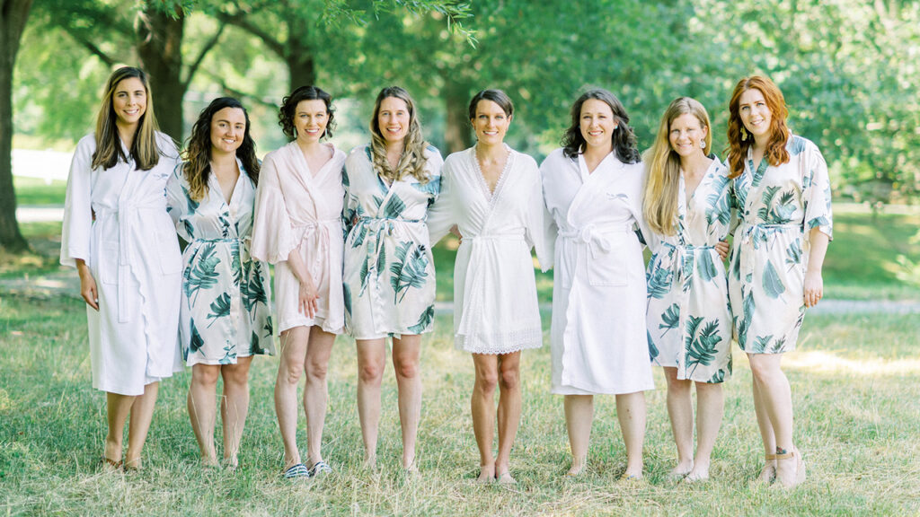 Seven women stand together outdoors on grass, smiling, wearing light-colored robes with floral or leaf patterns; trees and sunlight create a bright, natural background.