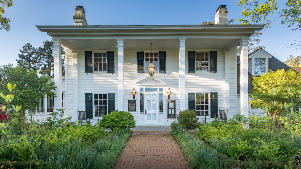 A white, two-story Southern-style house with columns, black shutters, a centered front door with sidelights, two rocking chairs on the porch, and a brick pathway surrounded by lush green garden.