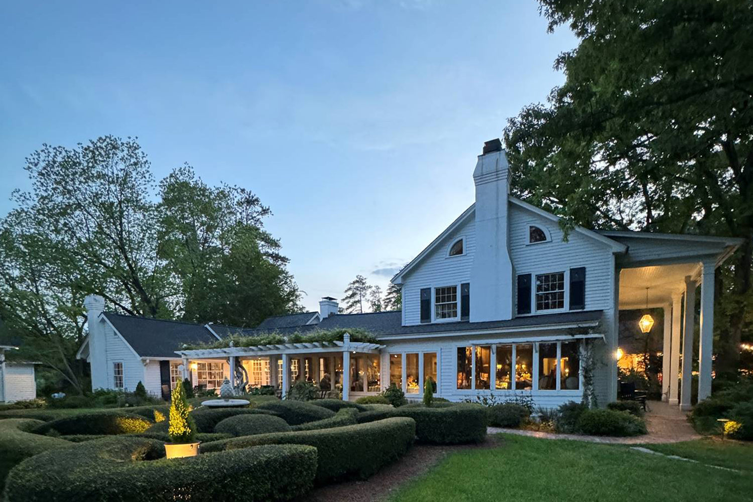 A large white house with tall columns and a chimney sits beside a manicured hedge garden at dusk, with warm lights glowing from inside the windows and trees surrounding the property.