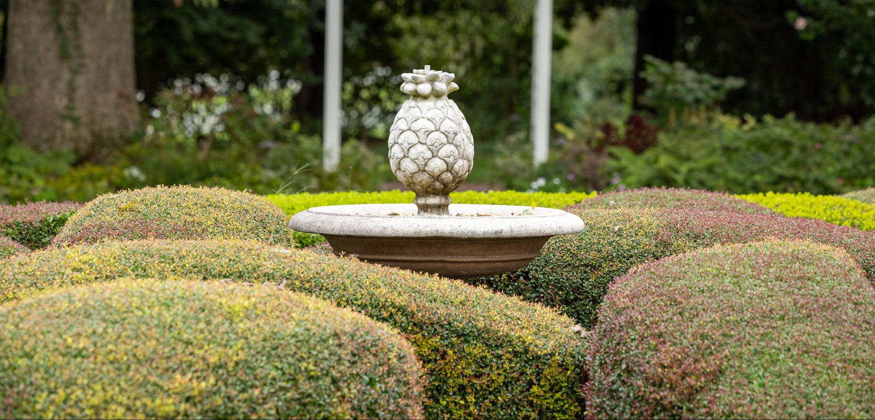 A white stone pineapple statue sits atop a circular pedestal, surrounded by neatly trimmed, rounded green bushes in a garden setting with various plants in the background.