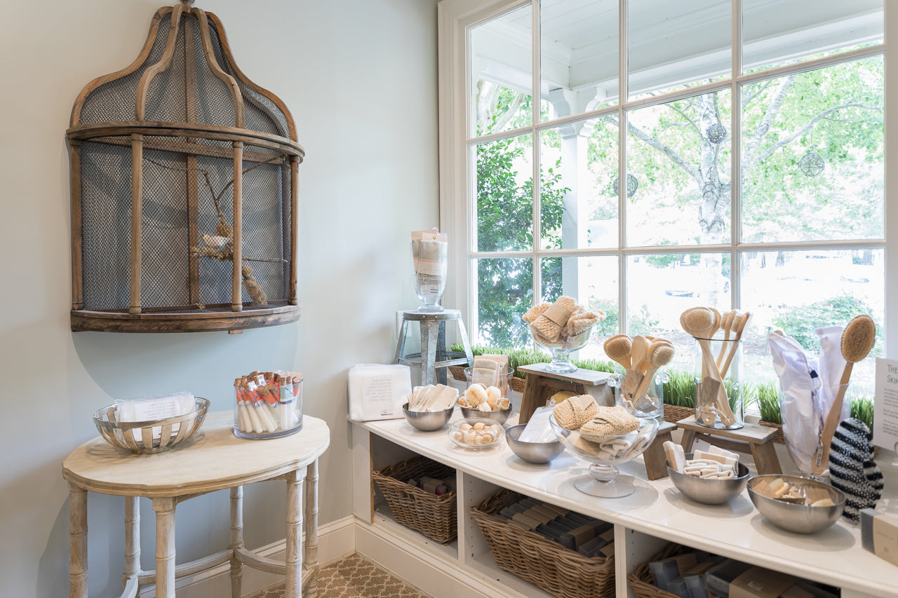 A bright boutique display with wooden shelves and baskets filled with natural bath products, brushes, and soaps, set by a large window with leafy views. A rustic birdcage hangs above a small table holding more items.