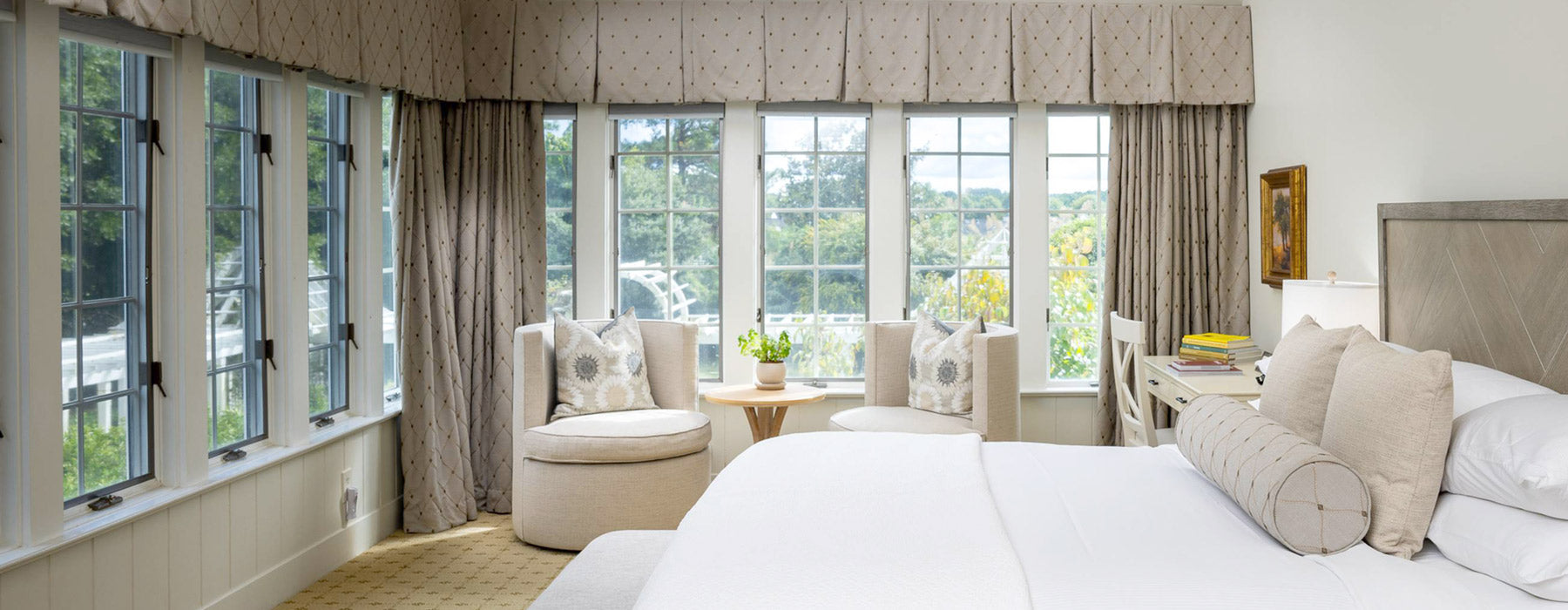A bright, elegant bedroom with large windows, two cushioned chairs around a small table, a desk with books, and a neatly made bed with white linens and neutral-toned pillows. Lush greenery is visible outside.