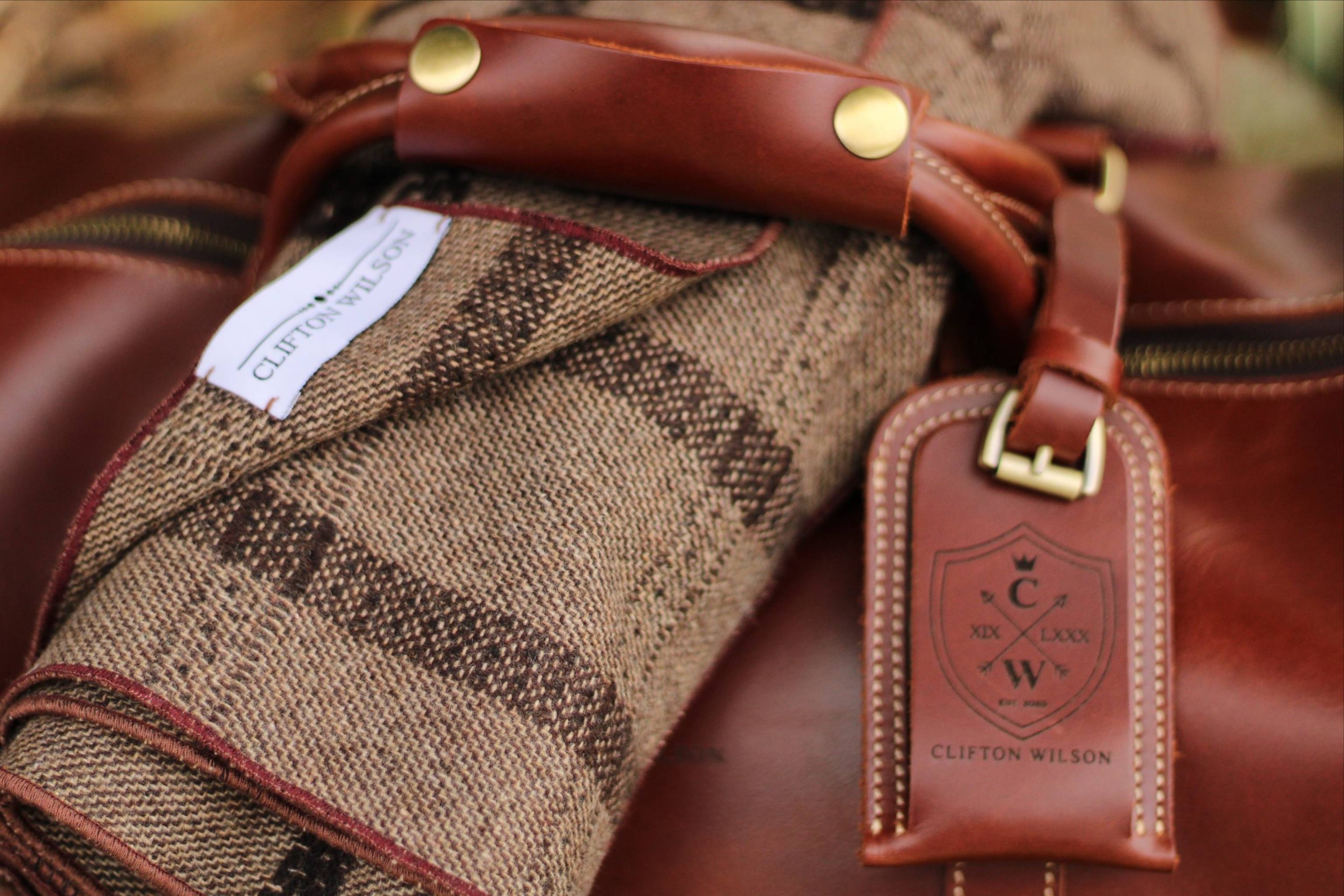 Close-up of a brown leather bag with gold hardware, a rolled-up woven blanket, and a leather tag featuring the Clifton Wilson logo and name. A fabric label with the same brand name is visible on the blanket.