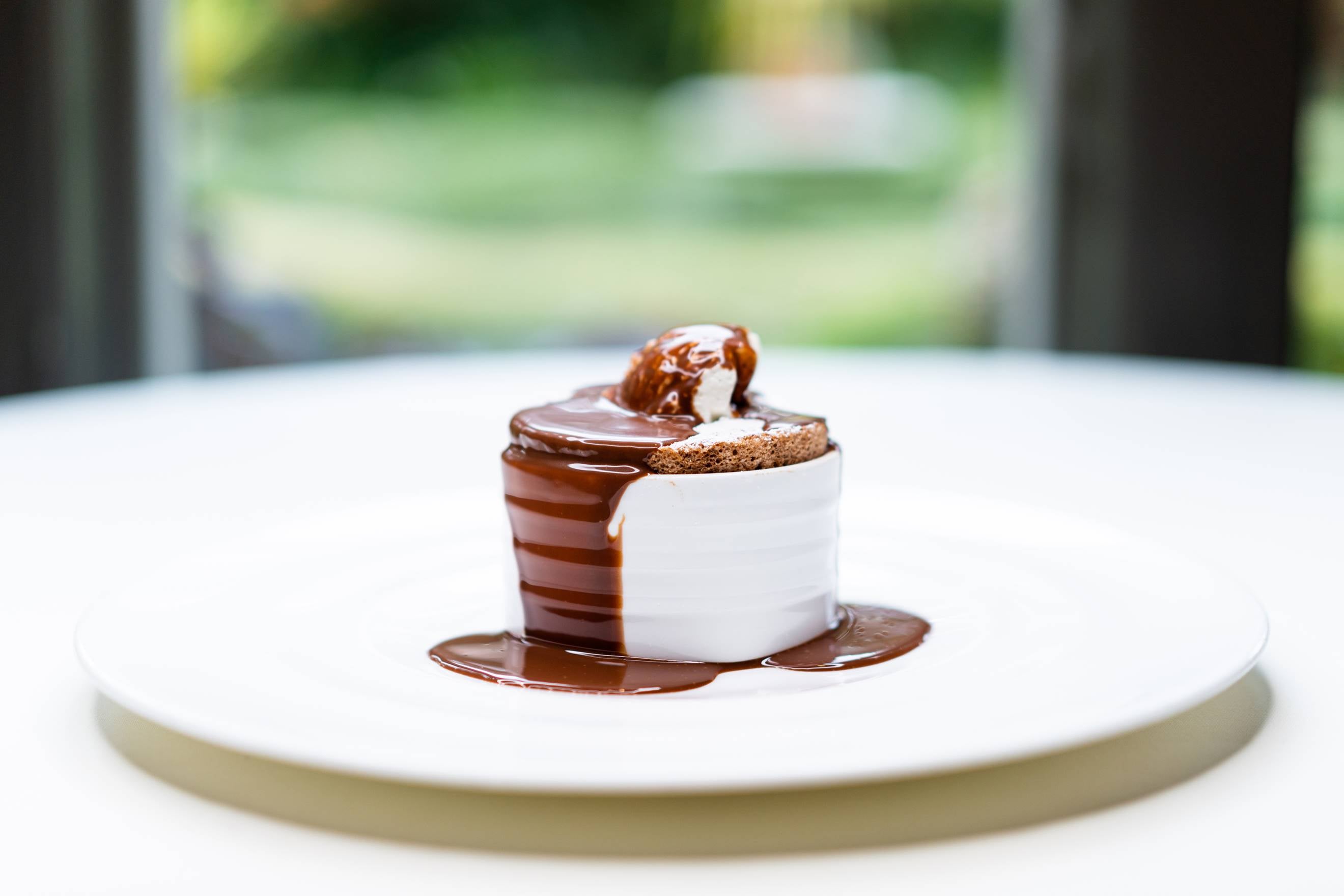 A chocolate soufflé in a white ramekin sits on a white plate, topped with a scoop of vanilla ice cream and drizzled with rich chocolate sauce, with a blurred outdoor background.
