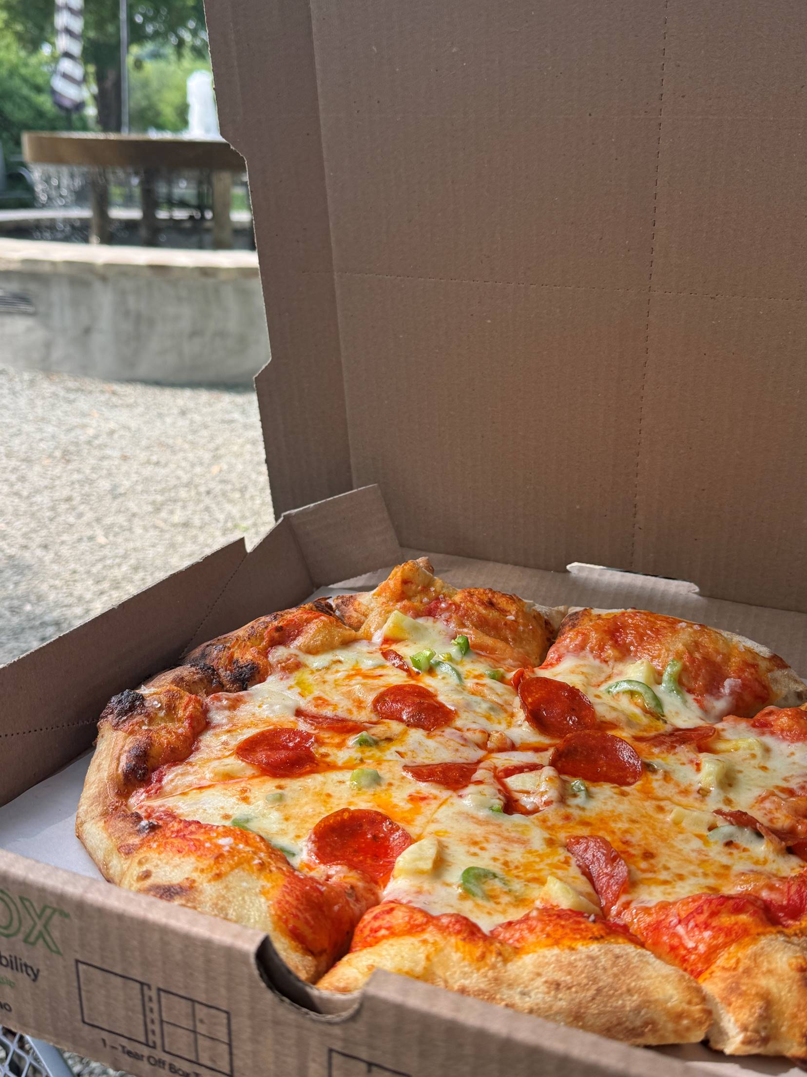 A freshly baked pepperoni pizza with melted cheese sits in an open cardboard box outside, with a picnic table and gravel area visible in the blurred background.