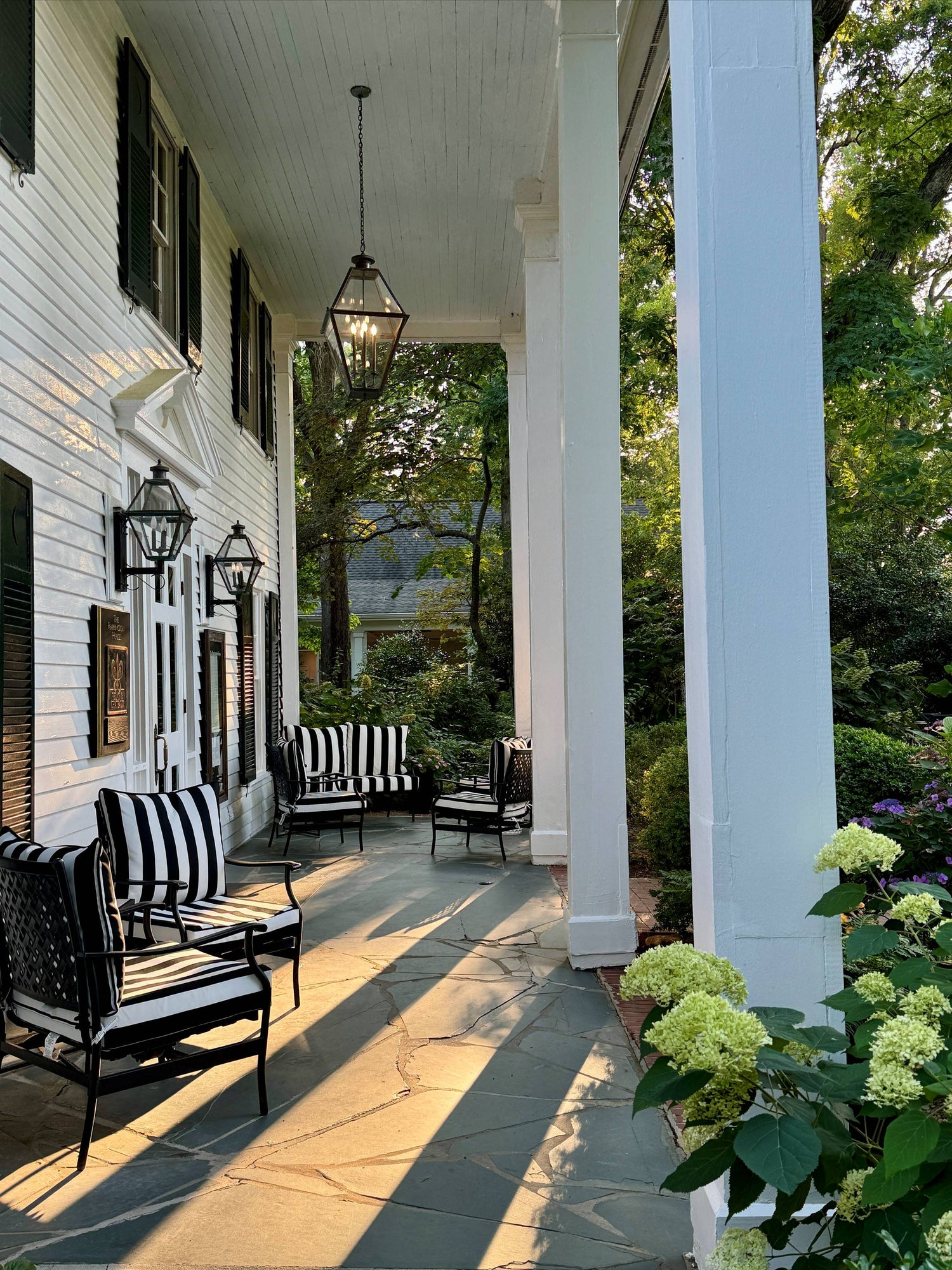 A sunlit porch with white columns, black-and-white striped chairs, lantern-style lights, and blooming hydrangeas, surrounded by leafy green trees and plants.
