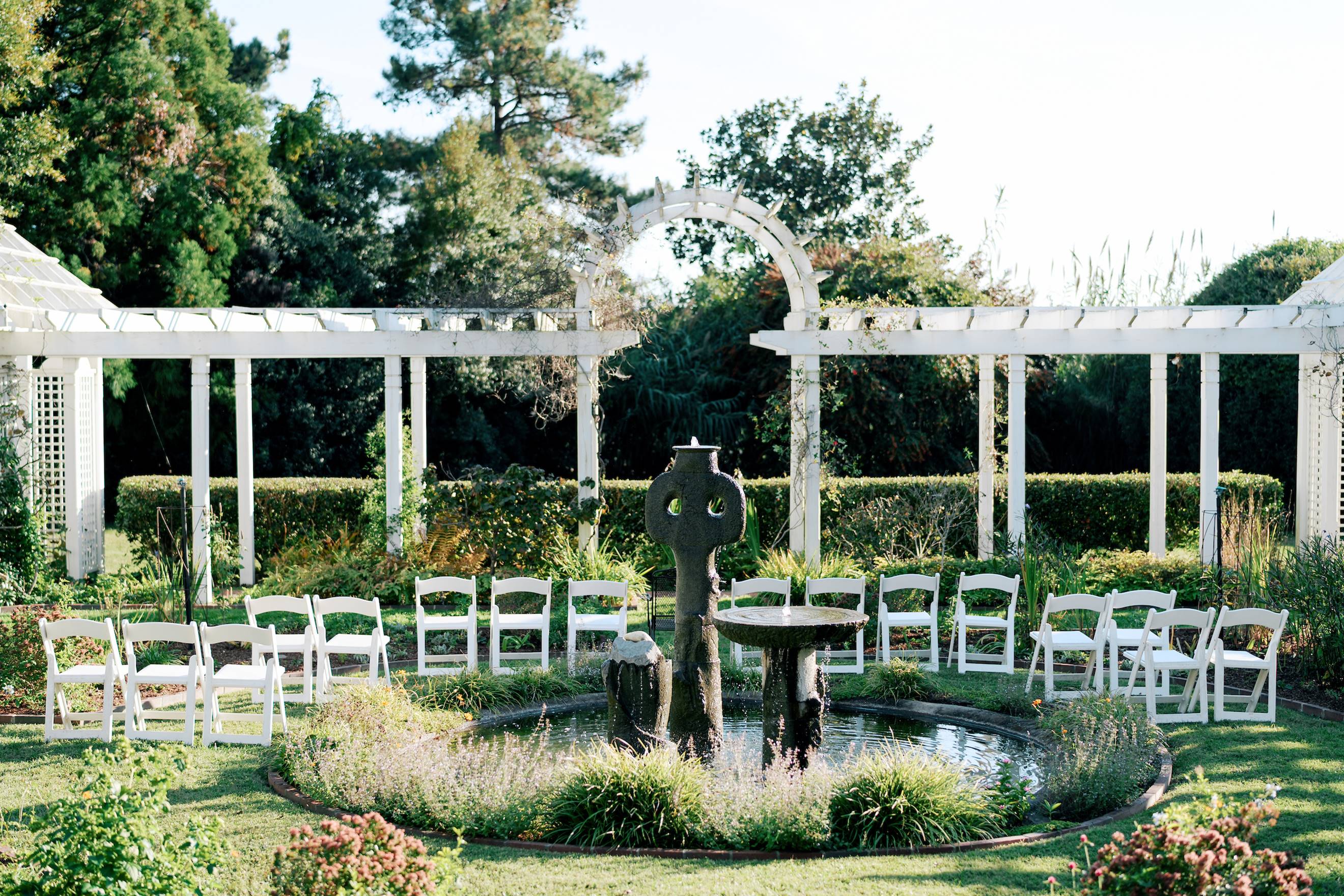 Rows of white chairs arranged in a semicircle around a stone fountain in a garden, surrounded by greenery, flowering plants, and white pergolas under a clear sky.