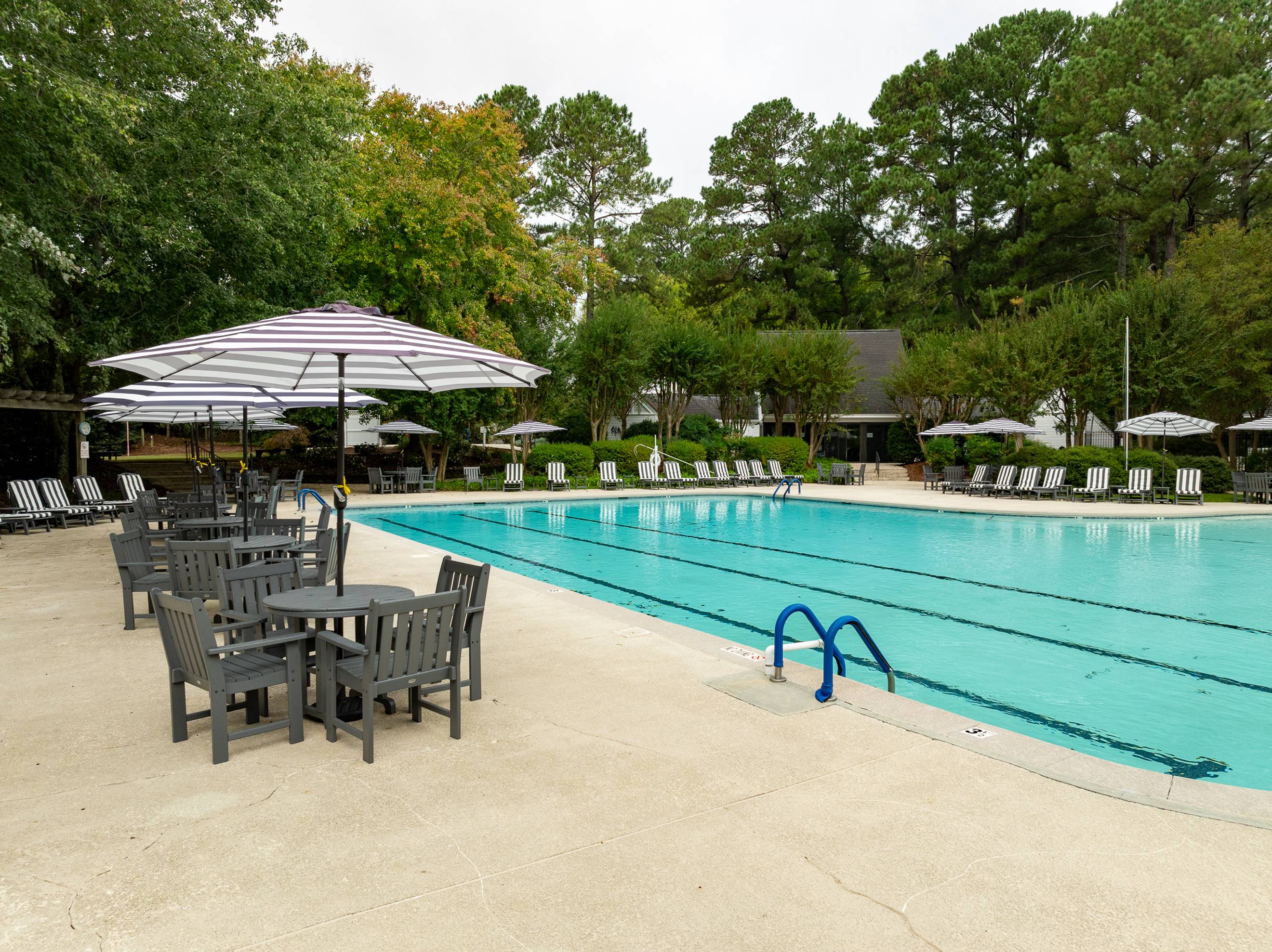 Outdoor swimming pool with clear lanes, surrounded by lounge chairs, tables, and umbrellas. Trees and greenery are in the background, and the pool area appears empty and peaceful.